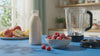 Milk, powder and raspberries being blended into a pink smoothie and poured into a glass on a blue counter.